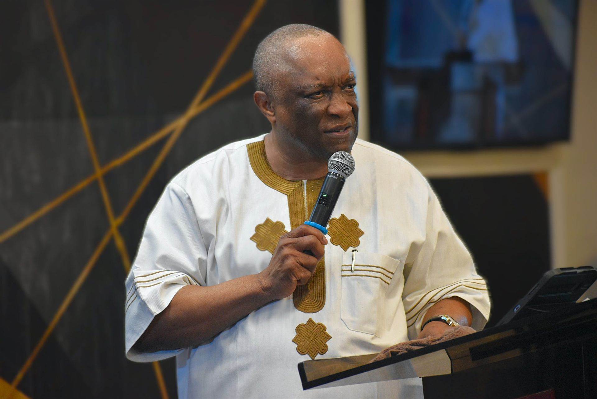 Man in white and gold shirt speaking into a microphone at a podium.