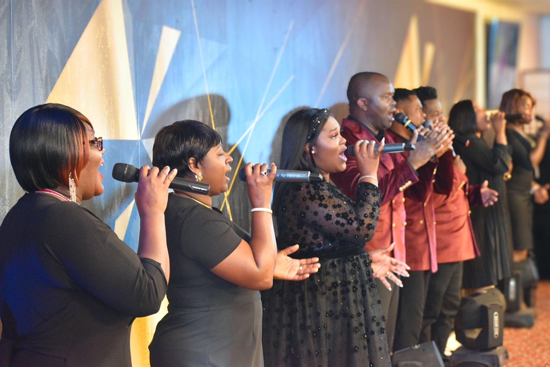 Choir singing on stage, some hold microphones, with bright background. People wear black, and red shirts.