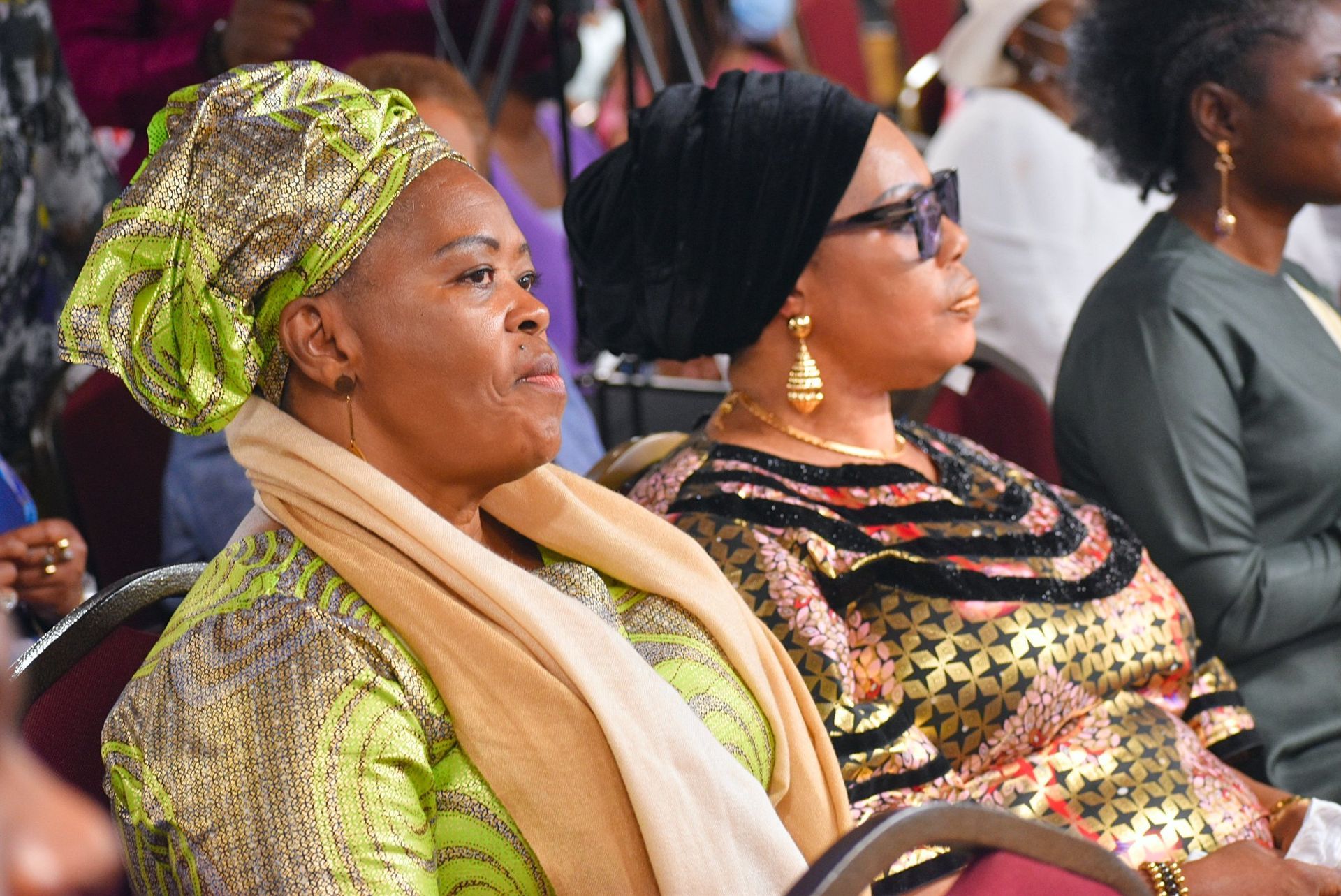 Two African women seated, one in green, the other in gold, attending an event, focused expressions.
