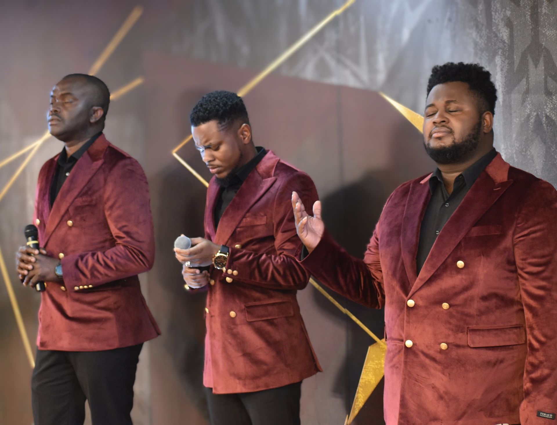 Three Black men in burgundy velvet blazers, singing on stage.