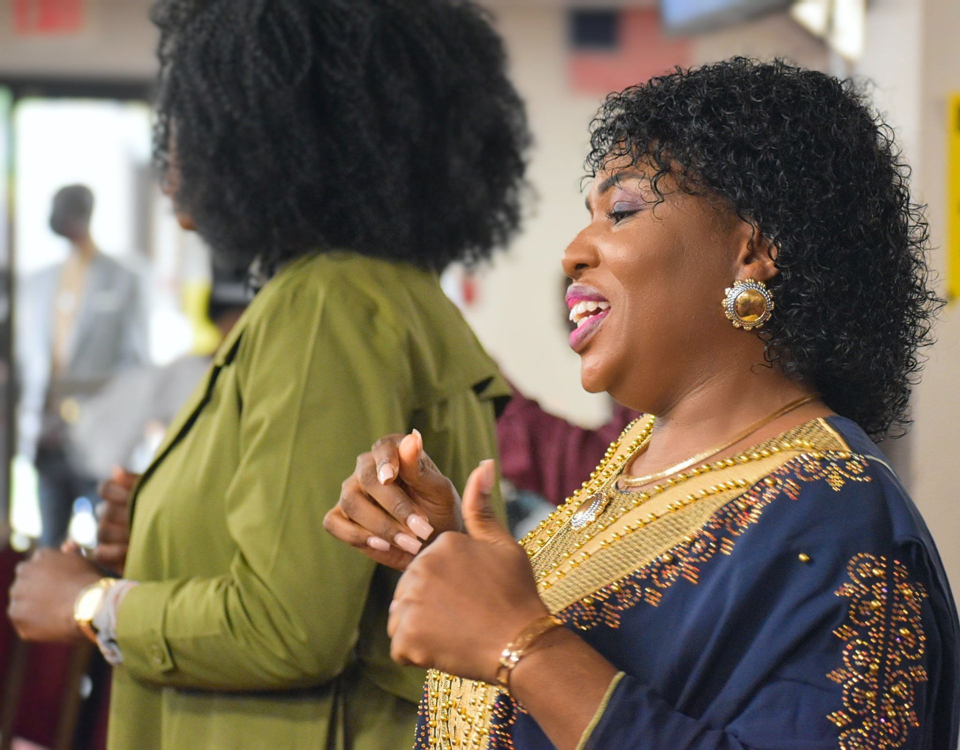 Woman in blue and gold dress sings, raises thumb, smiles; another in green jacket stands beside her.