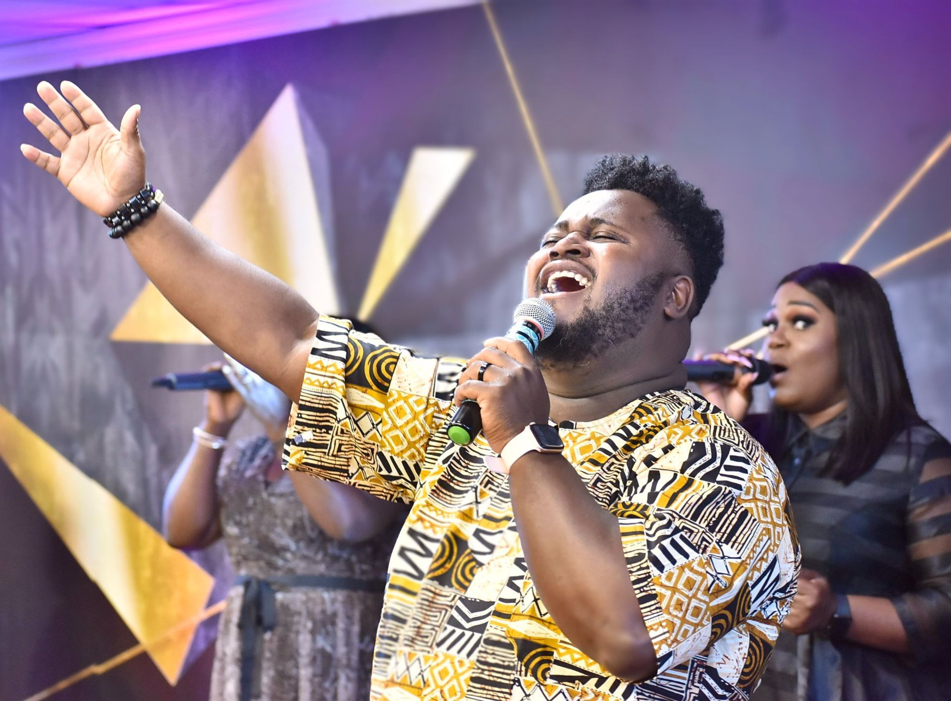 Man singing with arm raised, wearing patterned shirt; two women with microphones in background, stage.