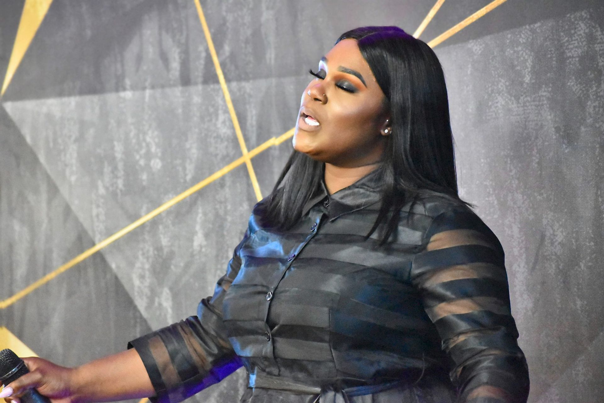 Woman singing into a microphone, wearing a black dress, in front of an abstract gold and gray wall.