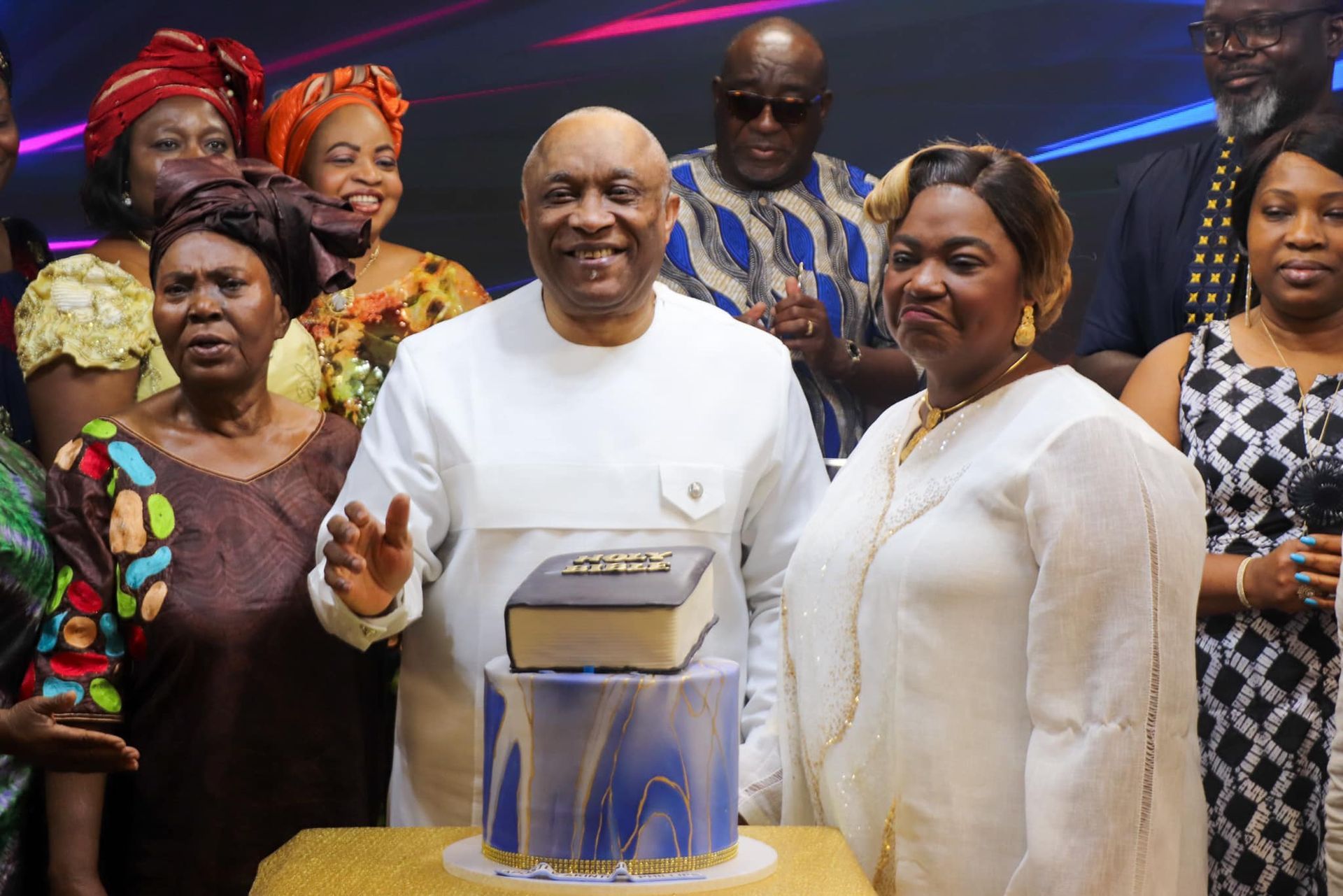 Group of people around a cake, smiling. Man in white, woman in gold, others in colorful attire.