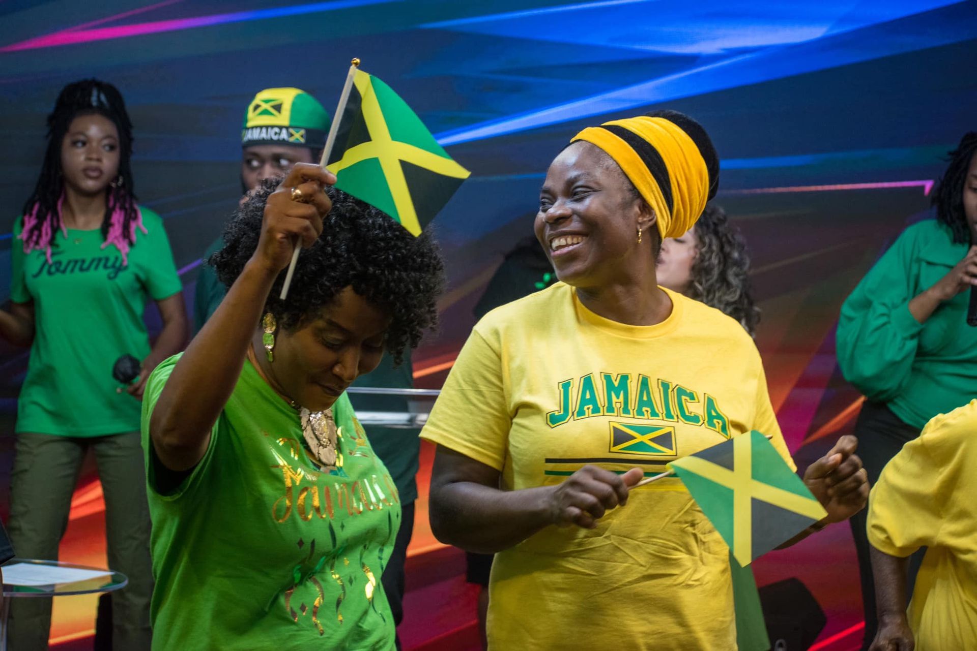 People celebrating, waving Jamaican flags. One woman smiles, wearing yellow shirt. Setting appears to be a stage.