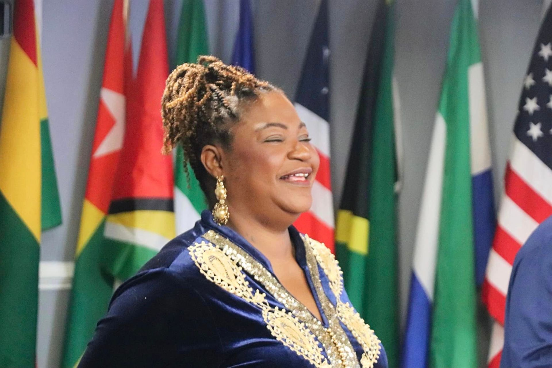 Woman smiling in a blue and gold embroidered dress, standing in front of various country flags.