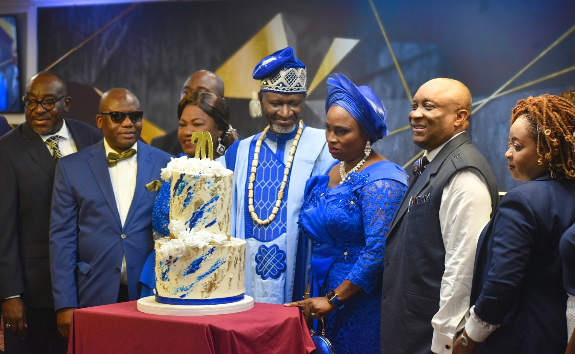 Group of people at a cake cutting ceremony. Man in traditional blue attire is cutting a cake.