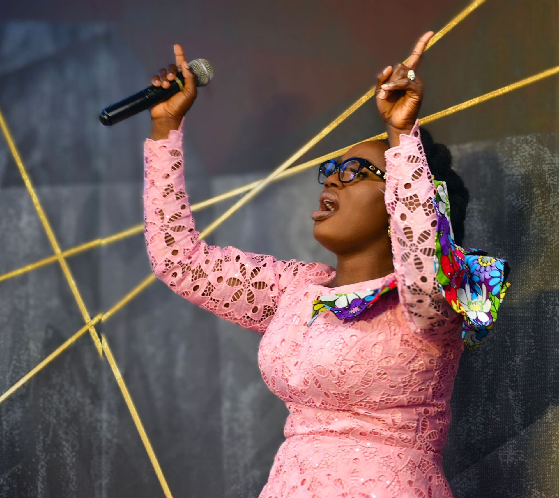 Woman with arms raised singing into microphone. Pink dress, glasses. Background with gold lines.