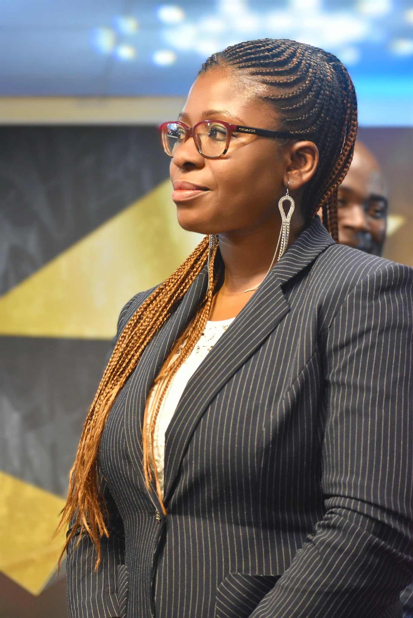 Woman in blazer, glasses, and braided hair looks off-camera. Another man partially visible in the background.