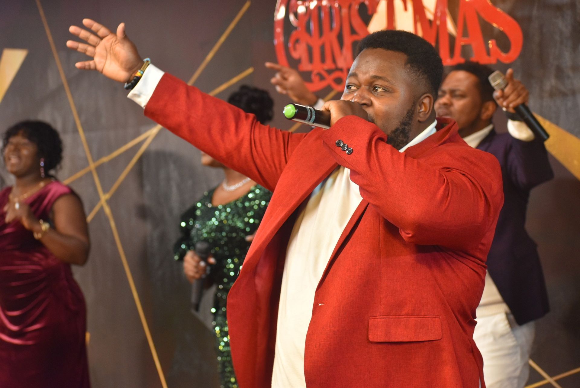 Man in red suit singing with group, Christmas backdrop.