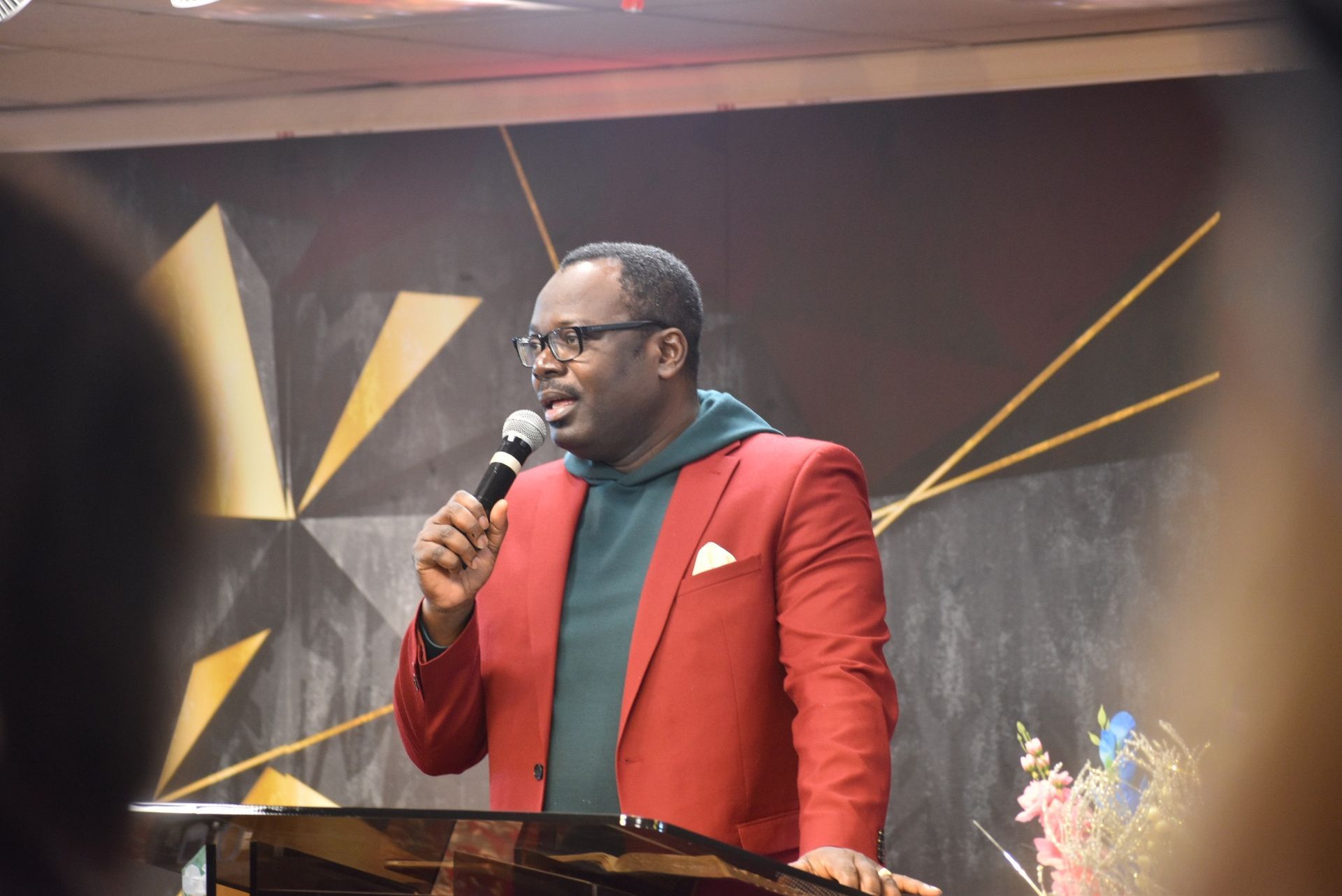 Man in red jacket speaking into a microphone at a podium, dark background with gold accents.