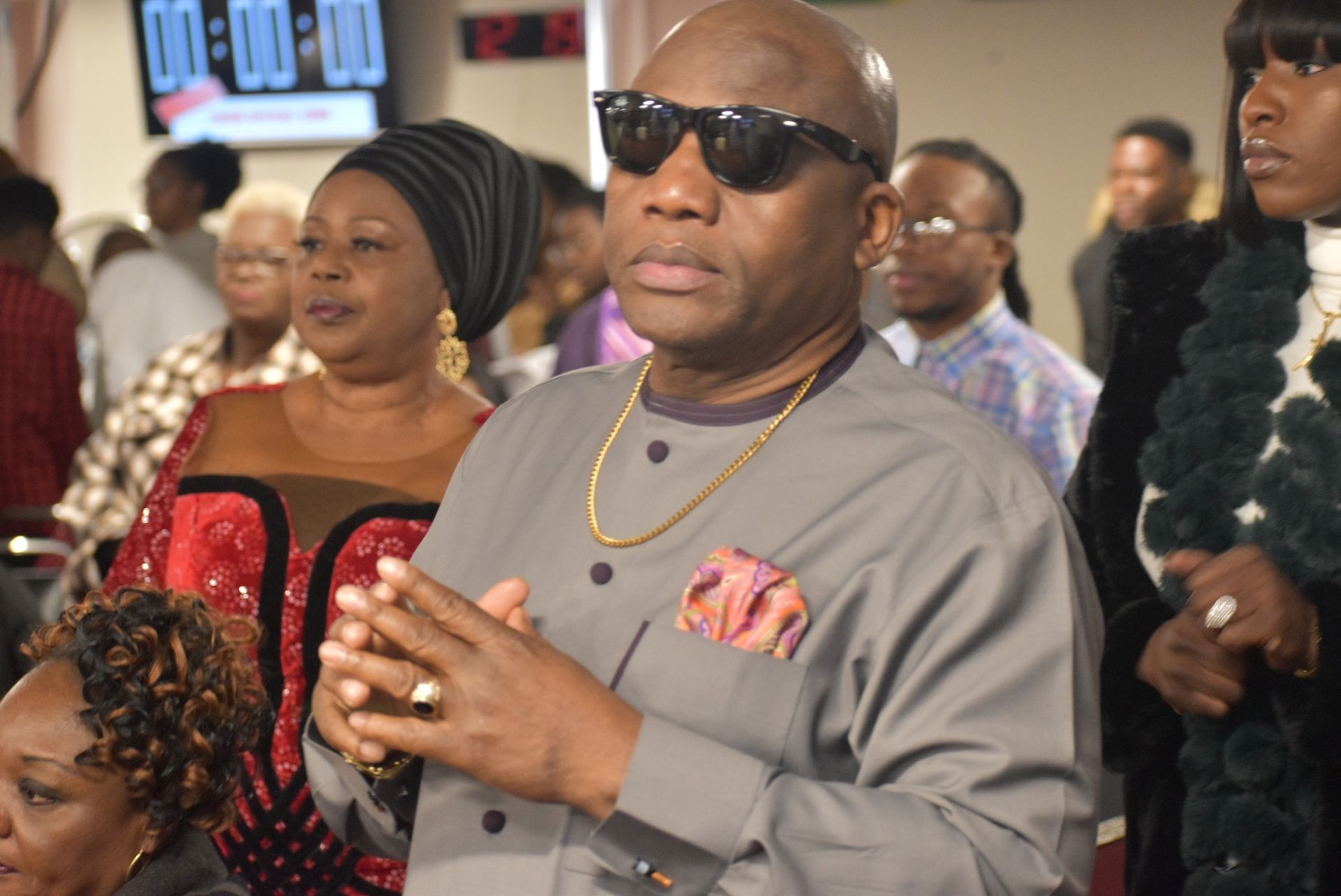 Man in gray shirt and sunglasses, with others in a room.