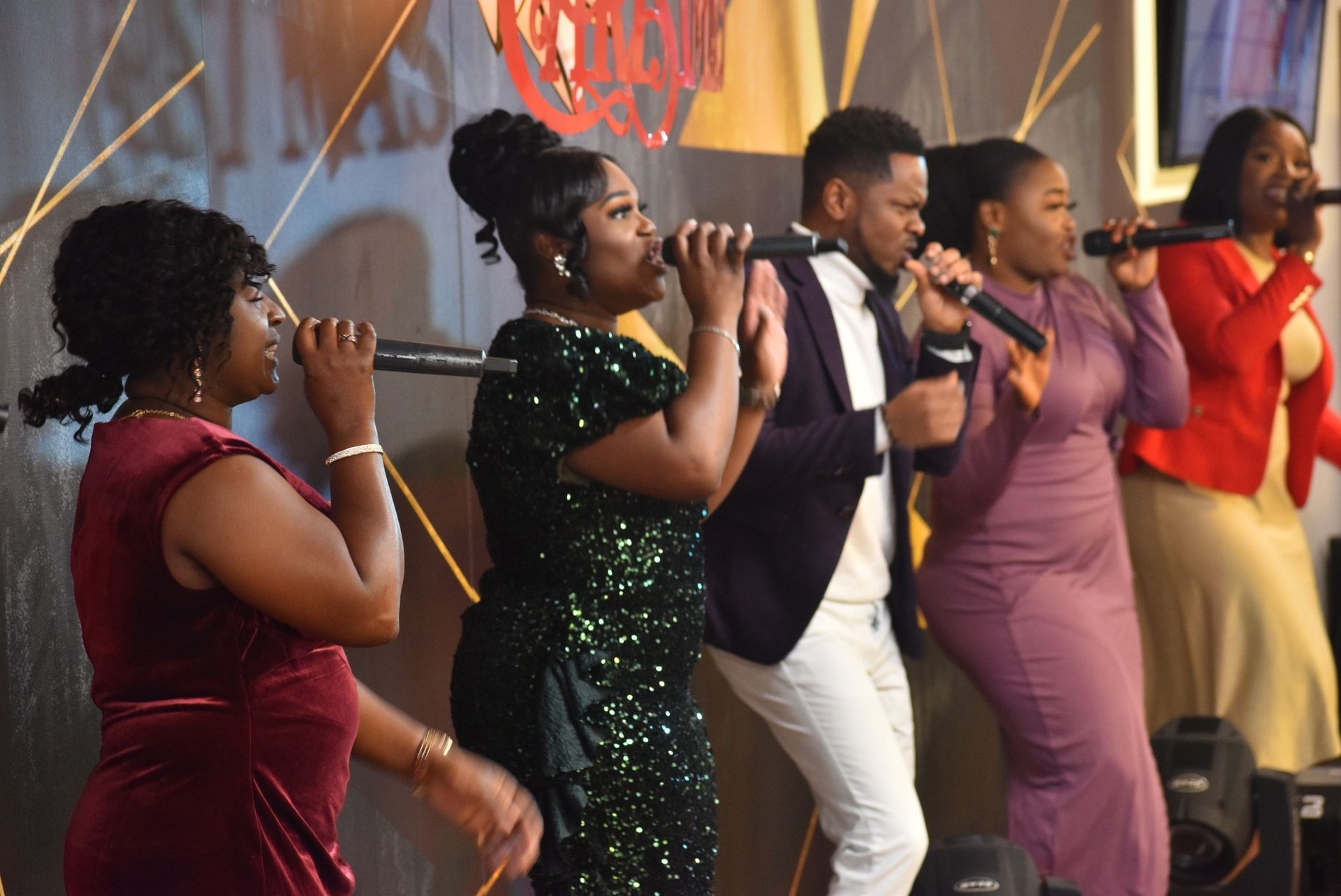 A diverse gospel choir singing with microphones. They are wearing colorful formal attire, in front of a stage.
