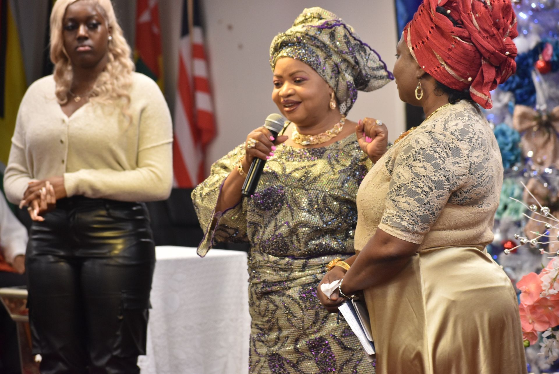 Woman speaking at event, wearing traditional dress, holding microphone, flanked by woman in gold and another in beige.