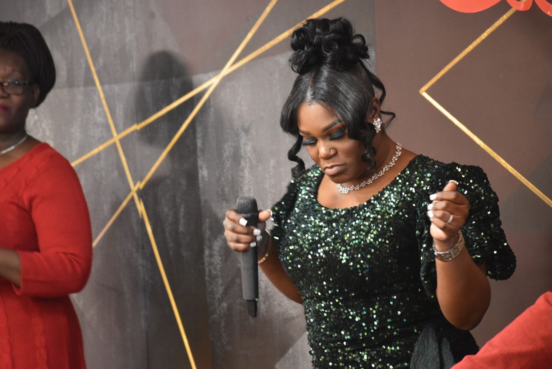 Woman singing into a microphone, wearing a green sequin dress. Background has gold geometric designs.