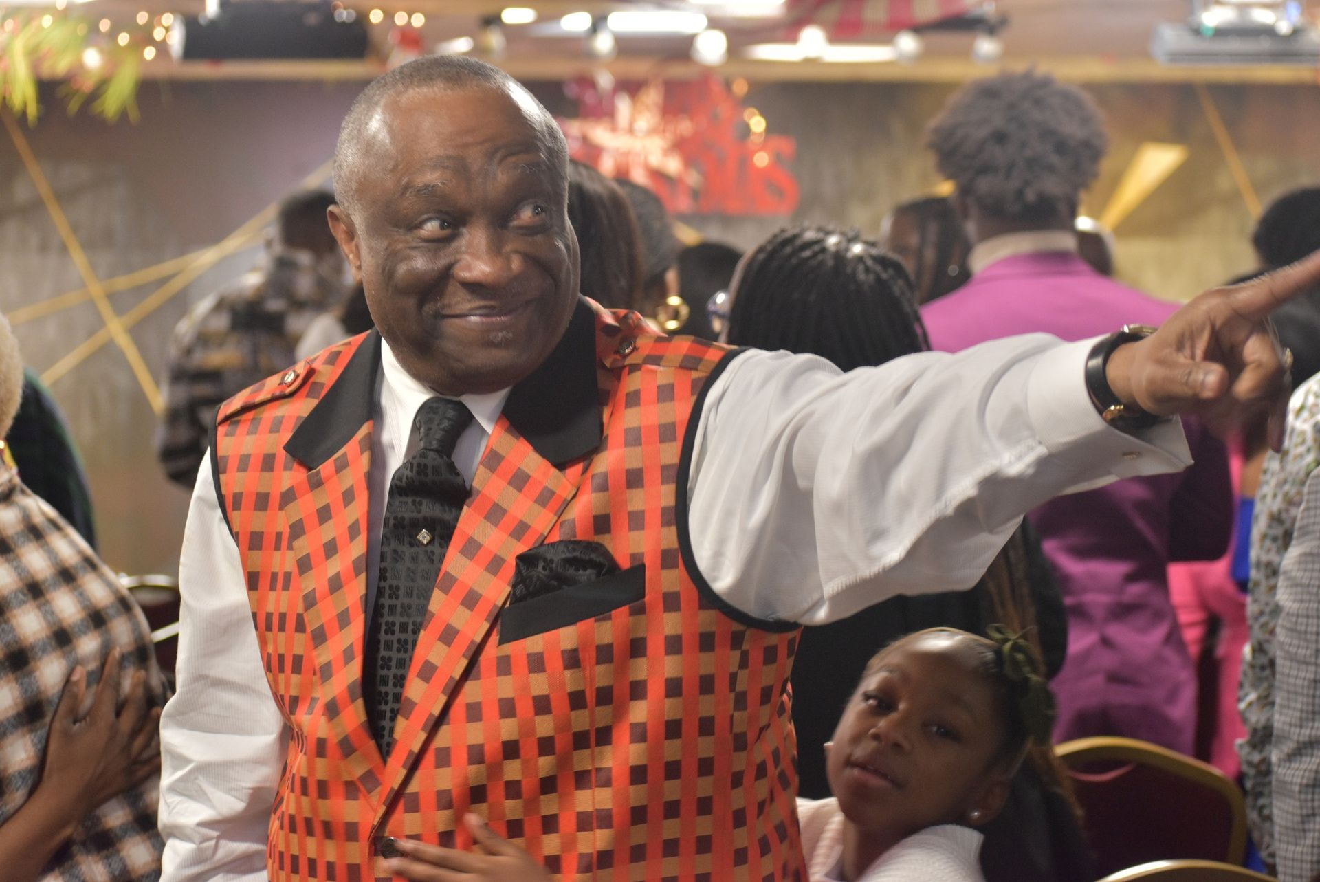 Man in orange checkered vest points, smiling in a crowded room, girl watches.