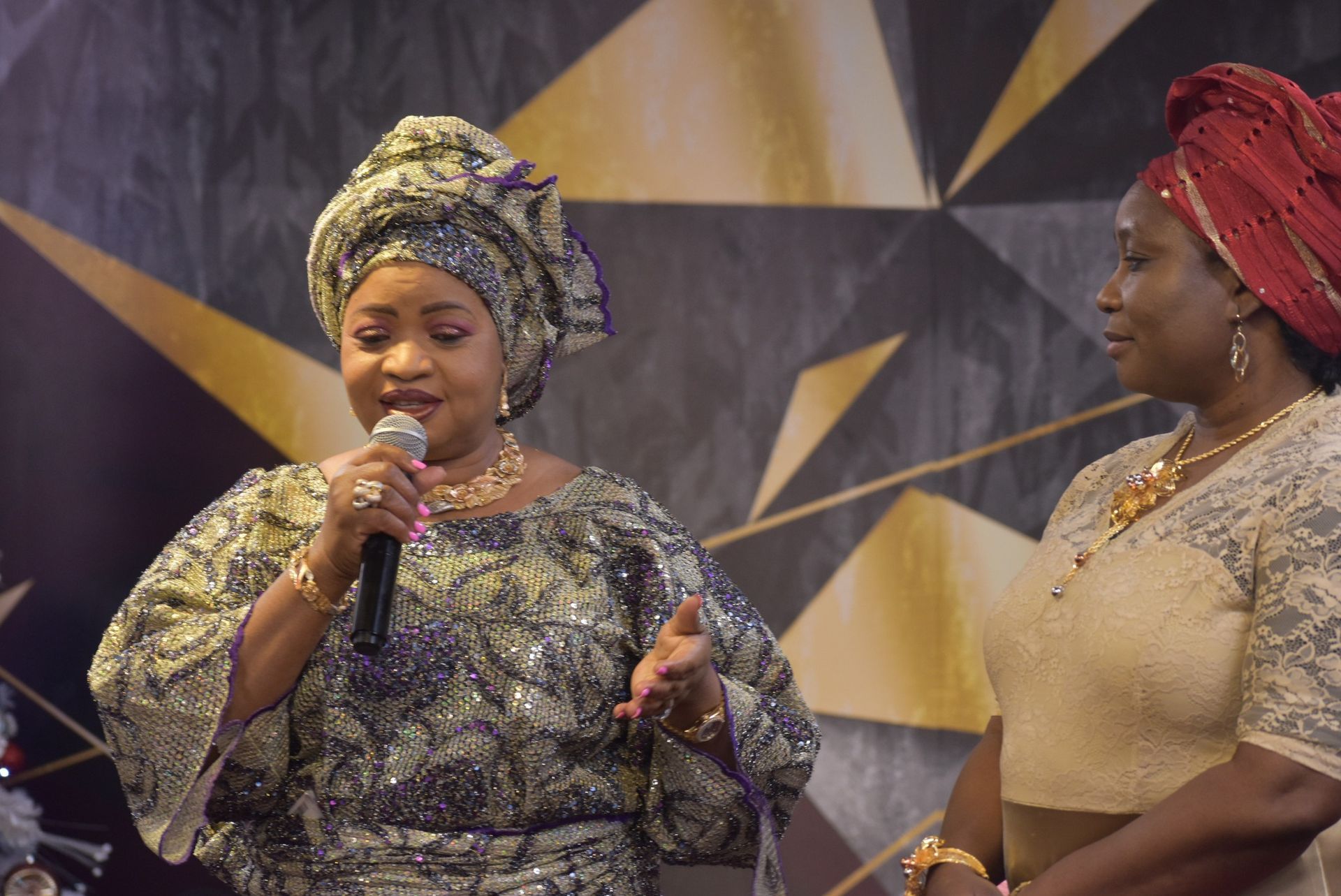 Two women in traditional attire on a stage. One speaks into a microphone; both smile.