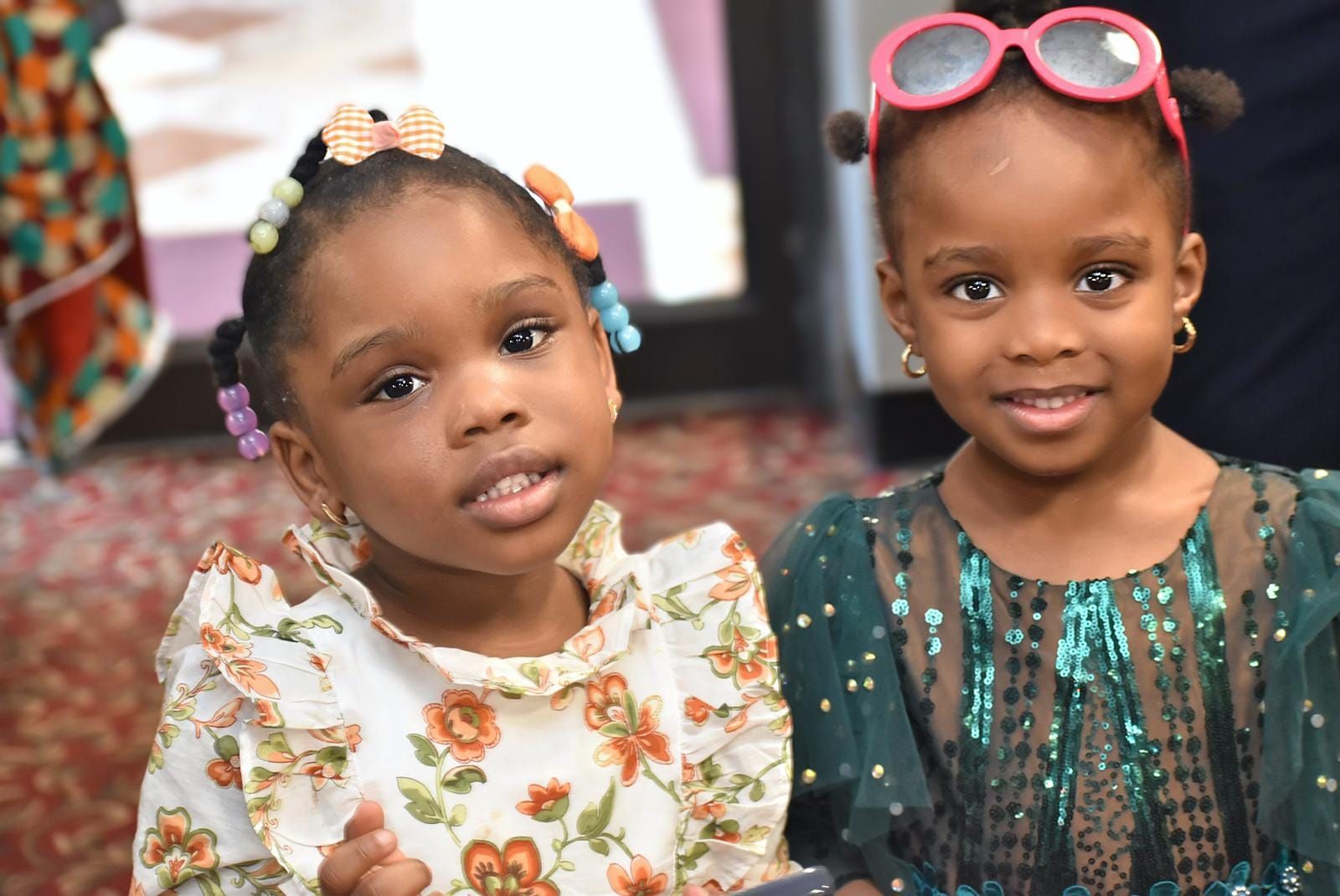 Two young Black girls smiling at the camera. One wears sunglasses on her head.