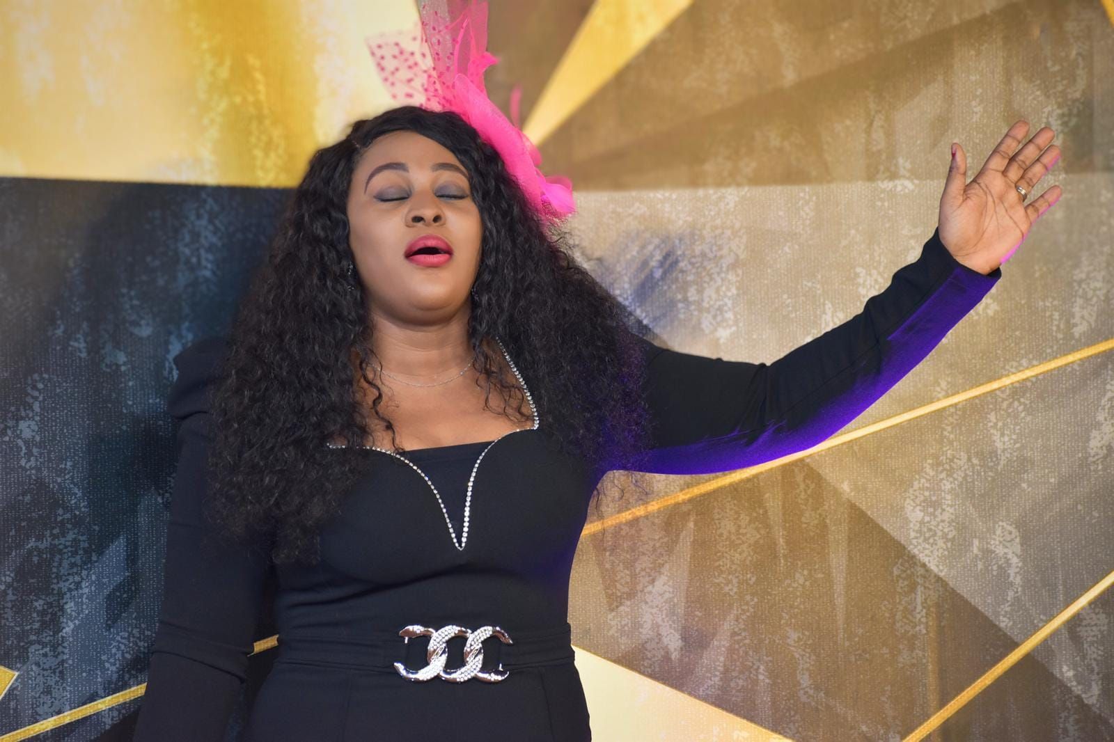 Woman in black dress with arm raised, eyes closed, singing in front of a patterned wall.