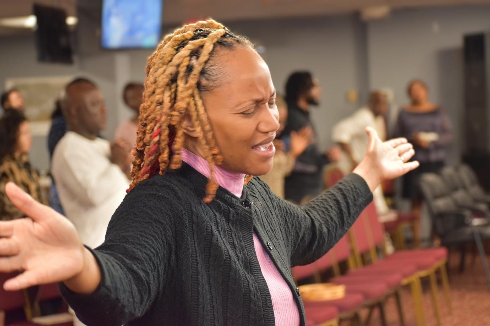 Woman with arms raised, eyes closed, in prayer, surrounded by people in a church setting.