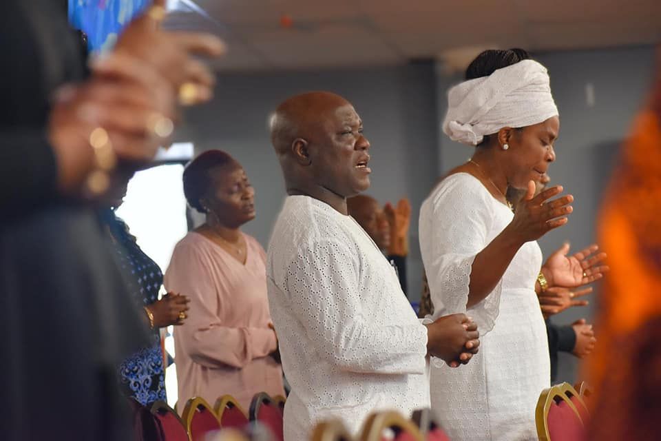 People praying in a church, some wearing white. Hands raised, focused expressions.