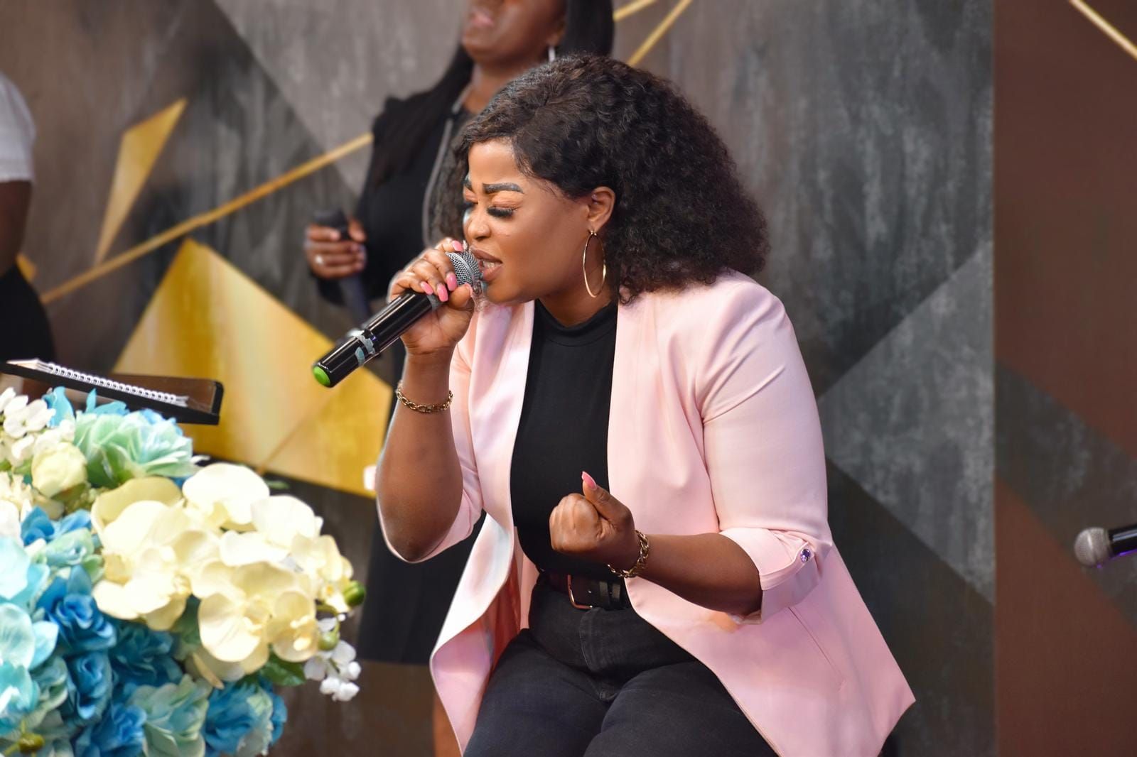 Woman singing into a microphone on stage, wearing pink blazer and black outfit.