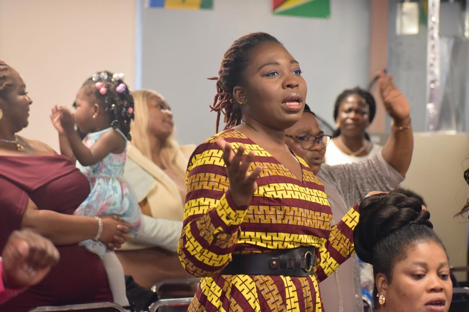 Woman in patterned dress leads worship with raised hands; people clap in a church setting.