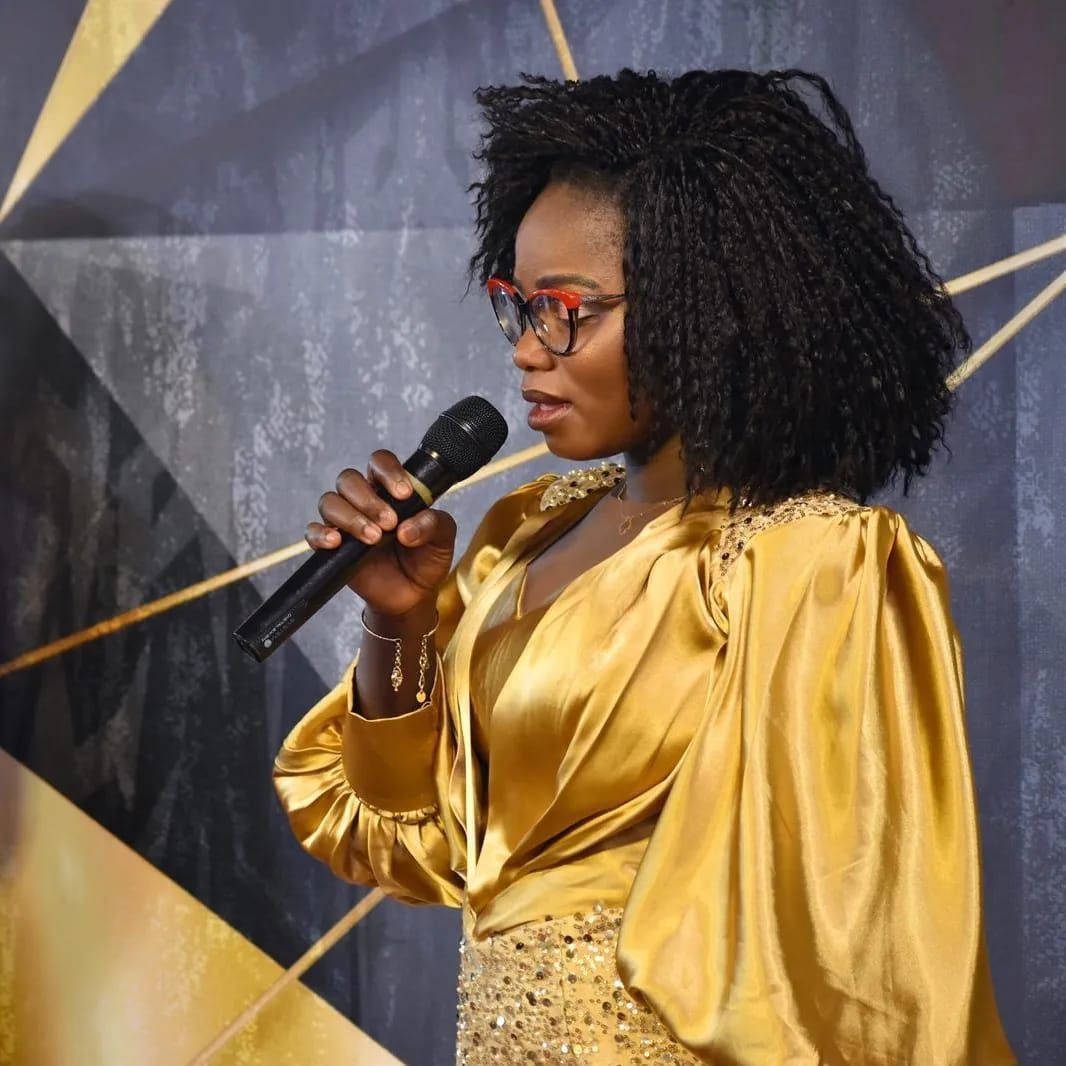 Woman with glasses in gold dress speaking into a microphone.