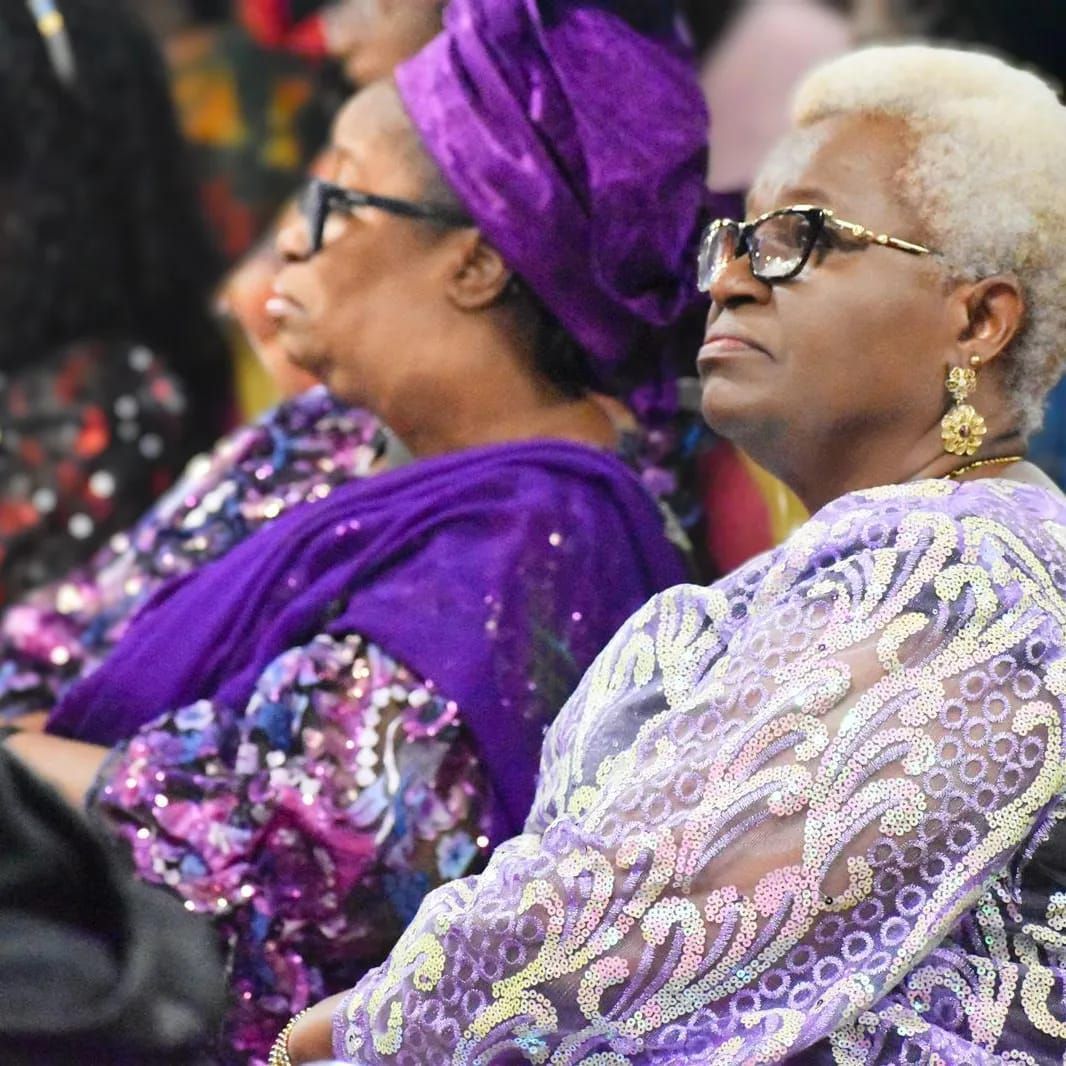 Two women in purple attire, wearing glasses, look forward, seated in a formal setting.