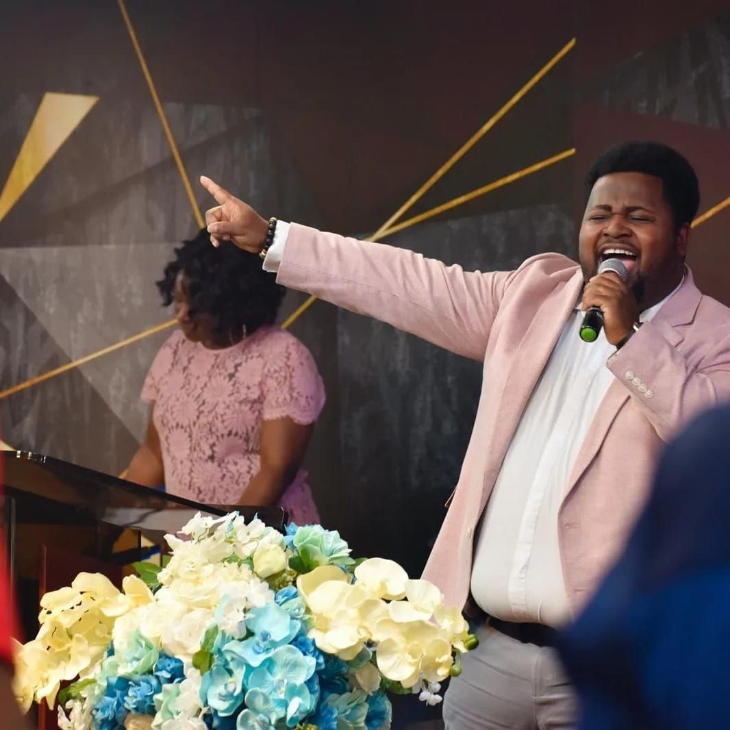 Man in pink blazer singing with open mouth, pointing upward, in front of a podium. Woman behind him, flowers below.