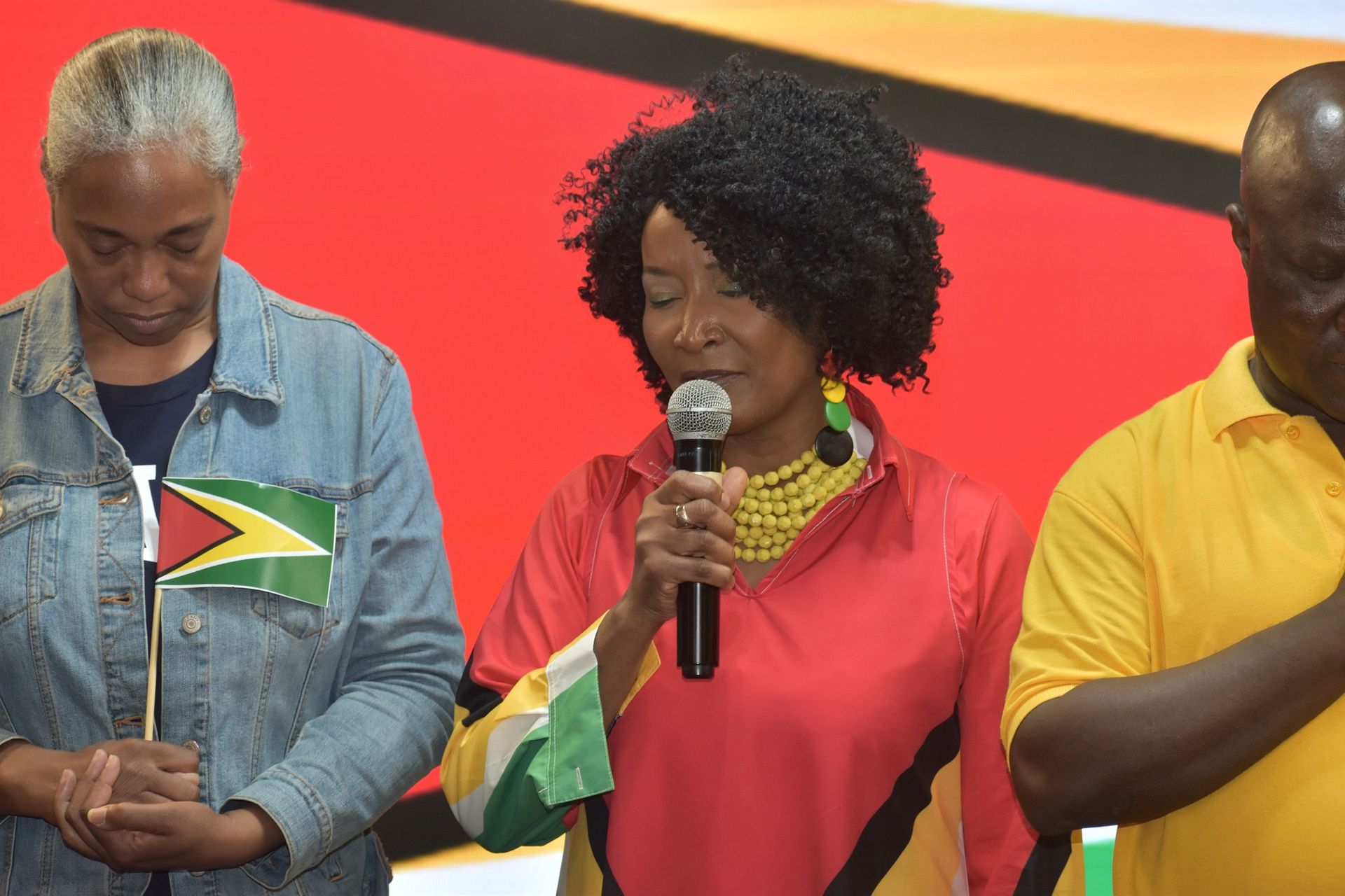 Three people, two women and a man, stand with heads bowed. The central woman speaks into a microphone.