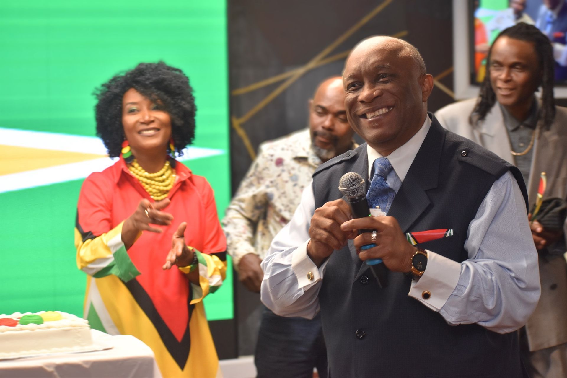People at event with cake. Man speaking into mic, woman gesturing. Guyana flag colors.