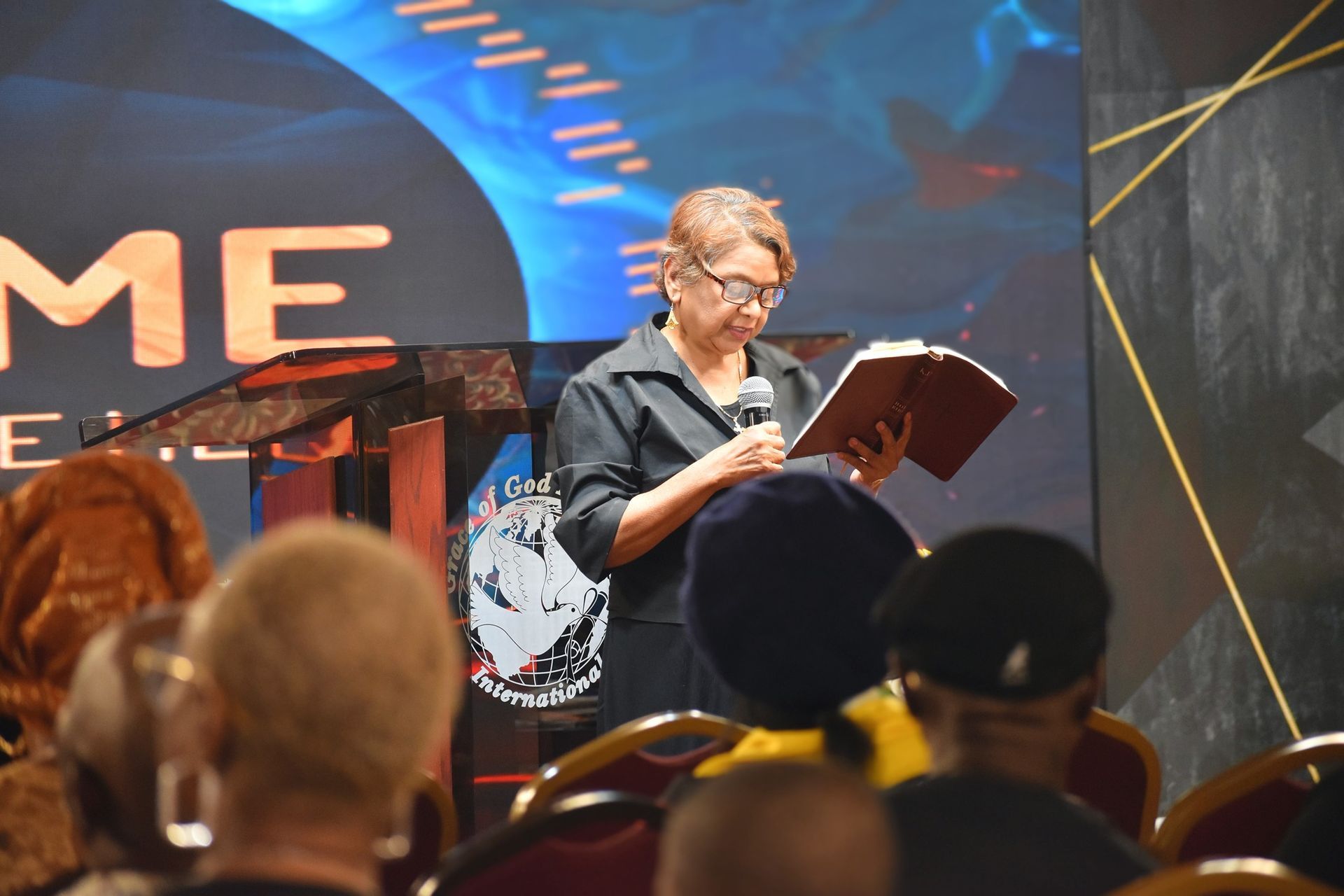 Woman in glasses reads from a book at a podium, speaking to an audience. Stage with 