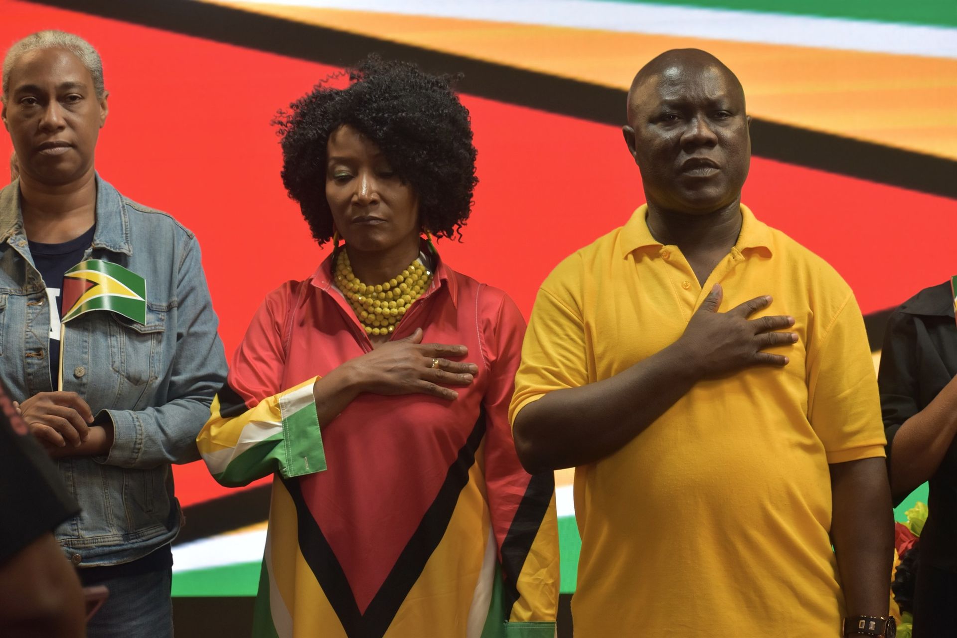 People with hands on chests, Guyana flag colors in background.