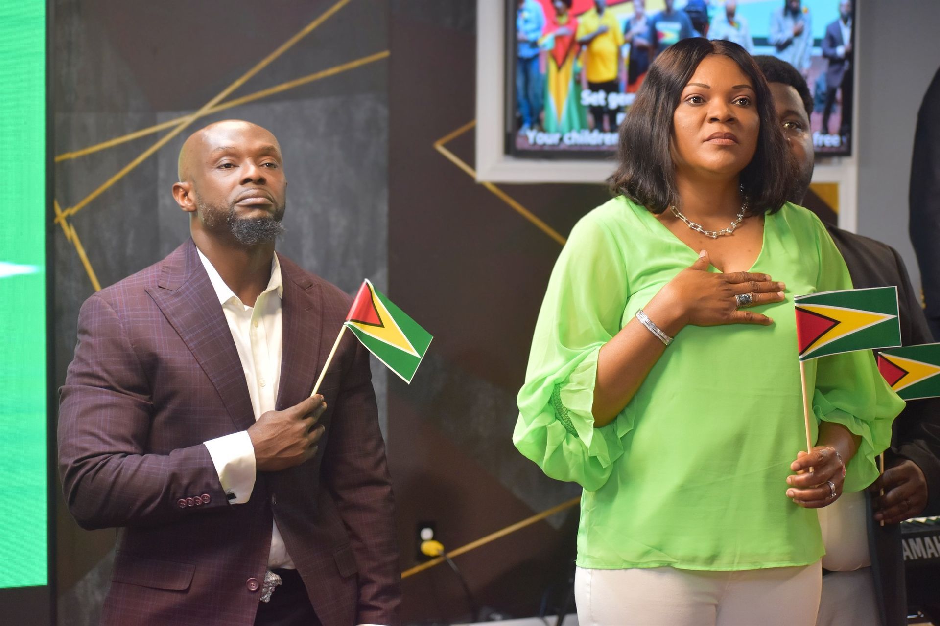 People with hands on chests, holding flags, in front of a screen. A man wears a suit. A woman wears green.