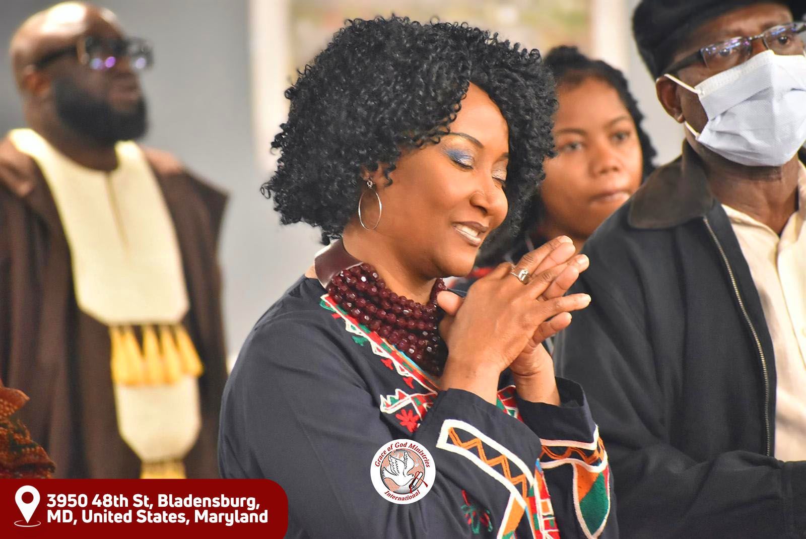Woman in traditional attire smiles, hands clasped. Others in background, Bladensburg, MD.
