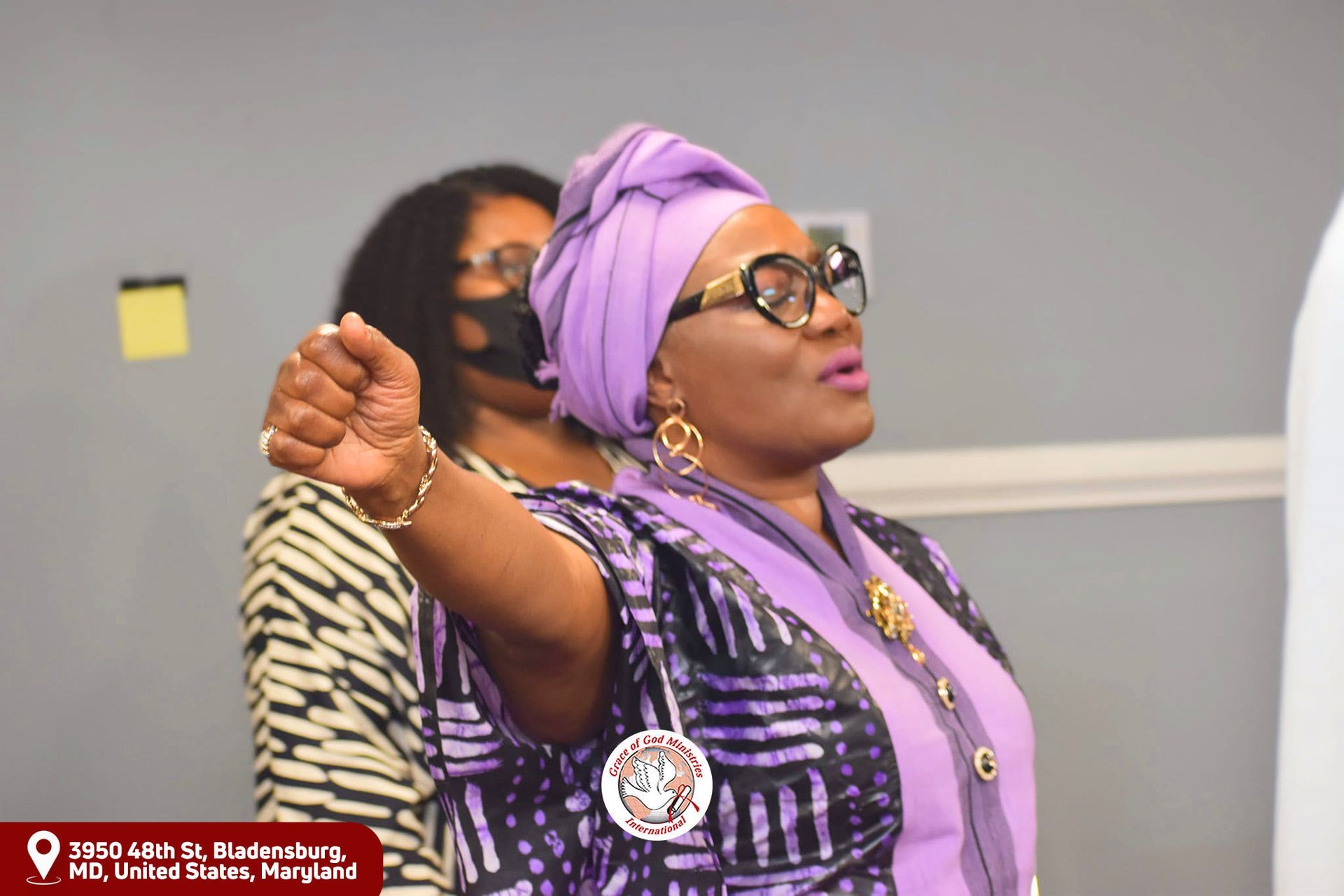 Woman in purple outfit raises fist.  Another woman with a mask in the background.