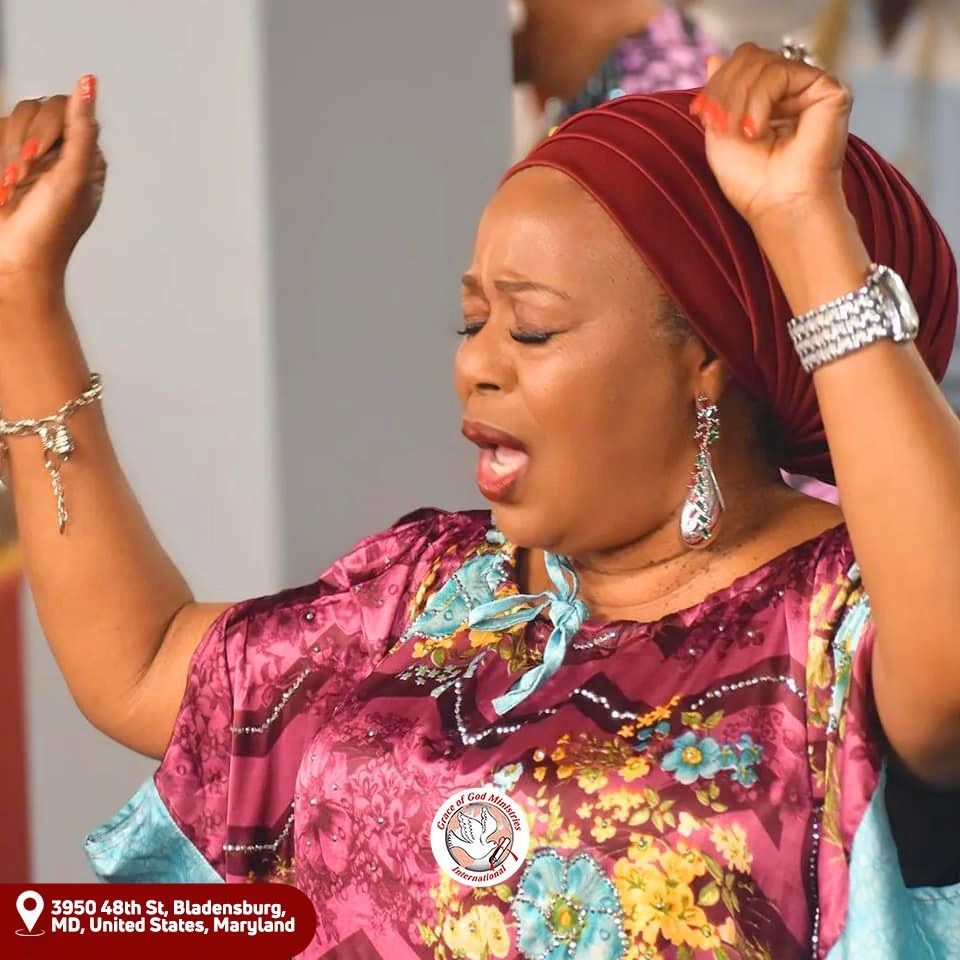 Woman in maroon headwrap and dress, arms raised in praise, eyes closed. Interior setting, church.