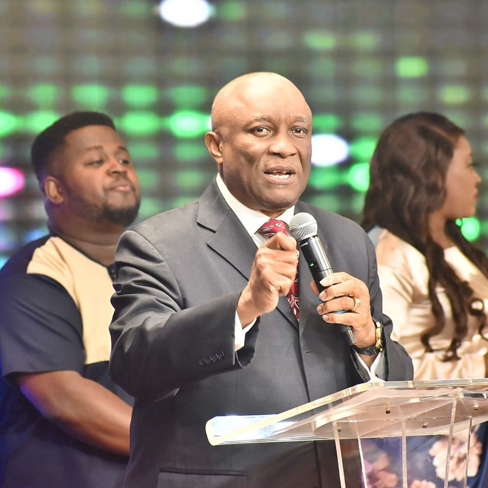 A bald man in a suit speaks into a microphone at a podium, with a small crowd behind him.