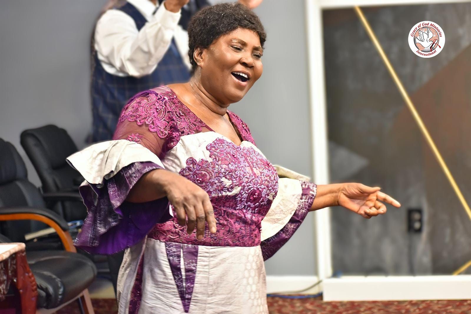 Woman in purple and white dress singing in a church setting.
