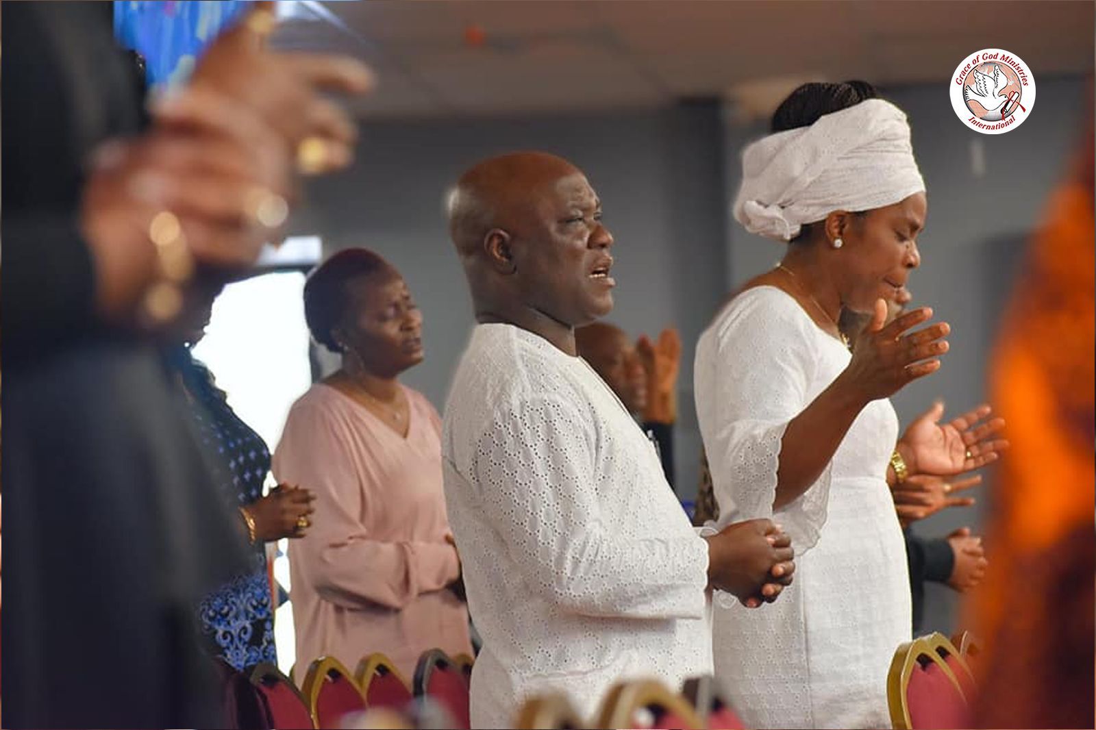 People in a church service, some in white, praying with hands raised.