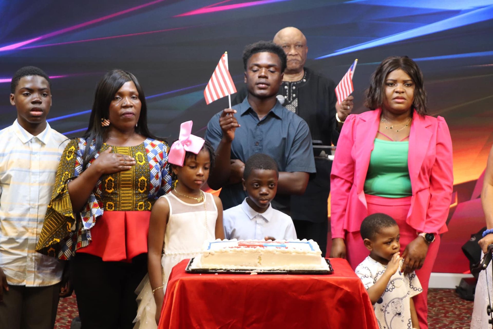 Family stands before a cake, some holding American flags, with hands on chests, celebrating.