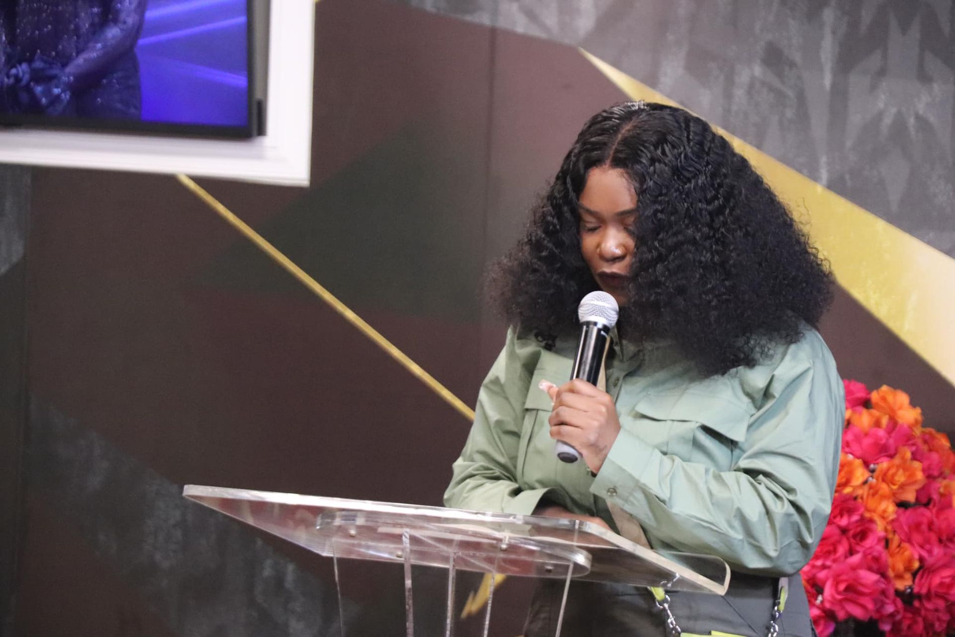Woman with curly hair speaks at a lectern, holding a microphone. She's wearing a green jacket. Flowers are nearby.