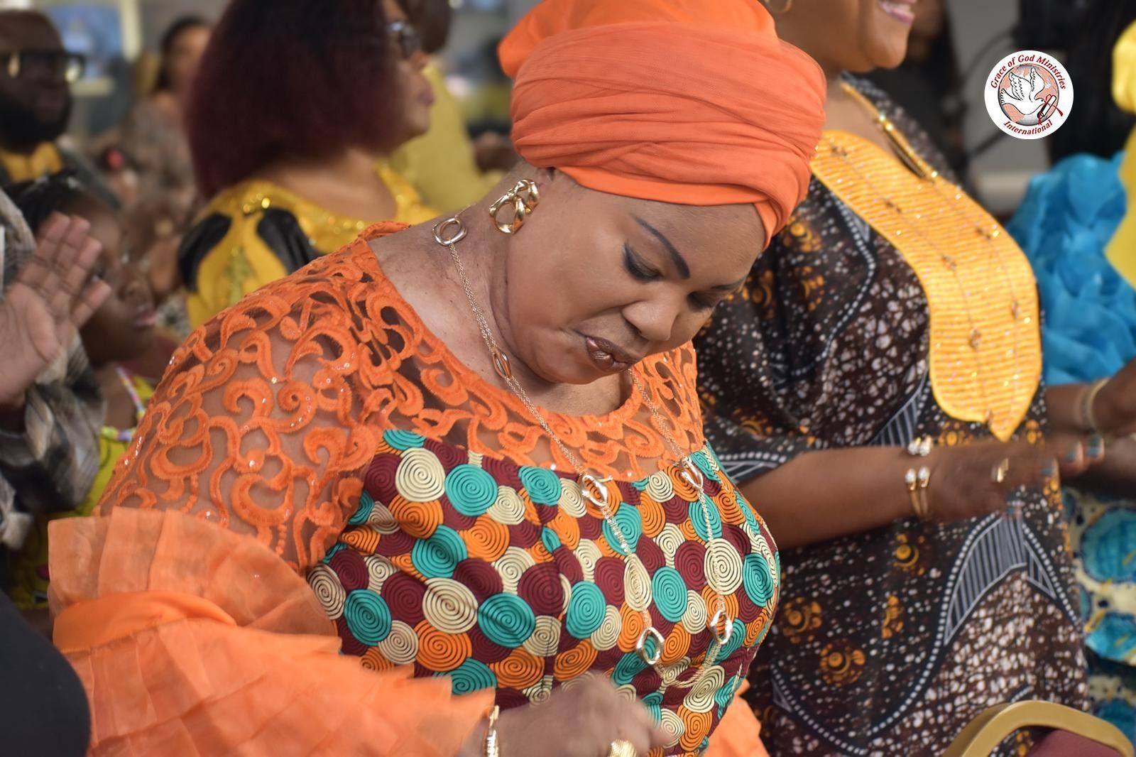 Woman in orange head wrap and lace top, eyes closed, possibly praying, at an event.