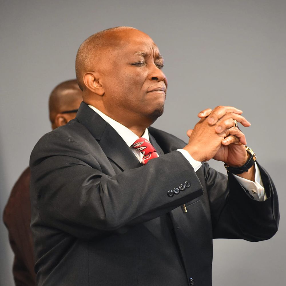 Man in suit with hands clasped in prayer, eyes closed, possibly in a church setting.