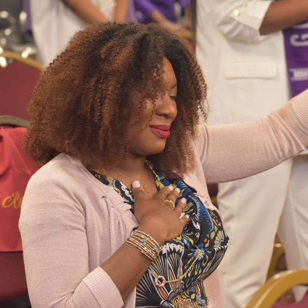 Woman with curly brown hair, hand on chest, smiles. Wearing floral top, pink cardigan.