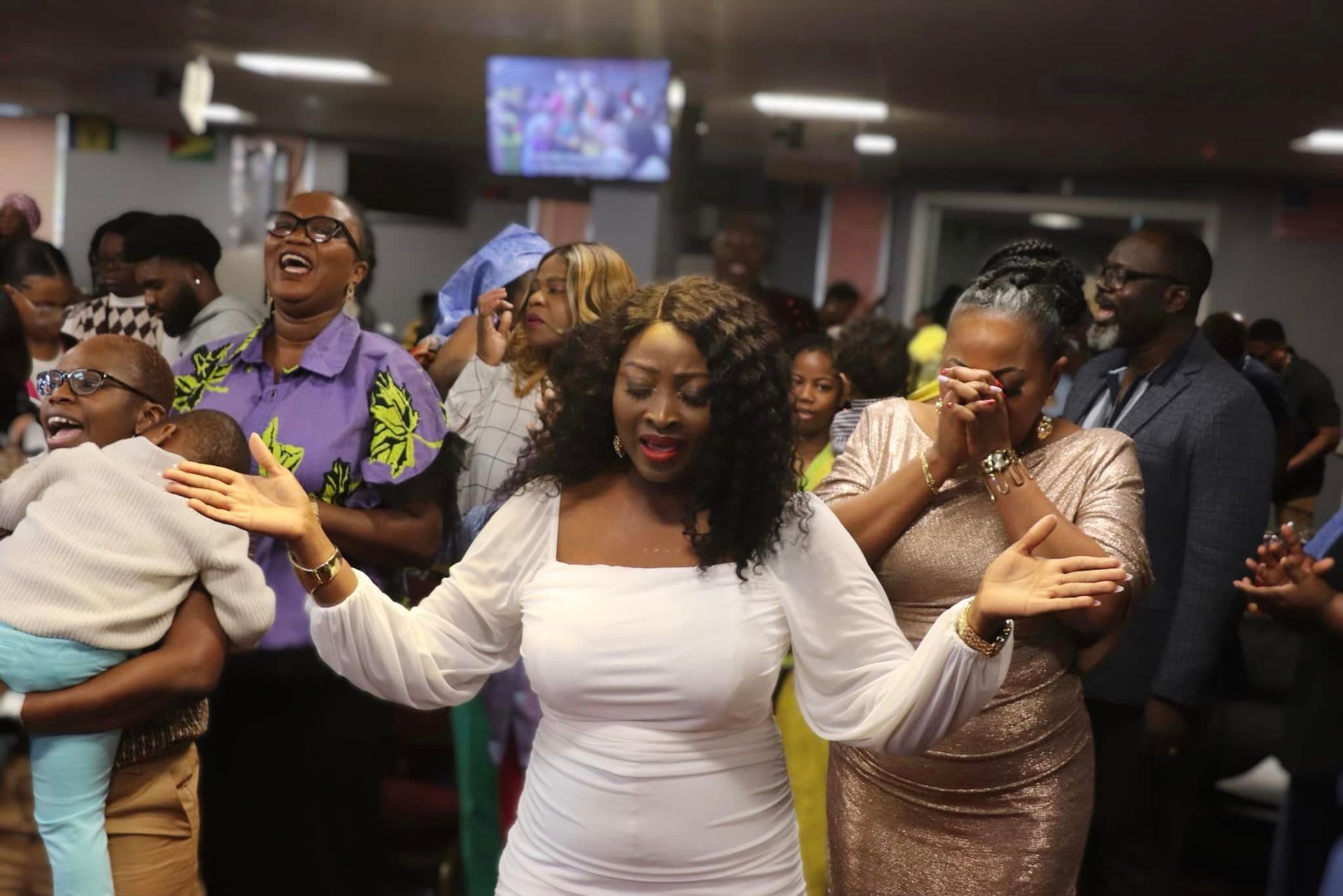 People in a church service; woman in white dress with arms raised, others singing and praying.