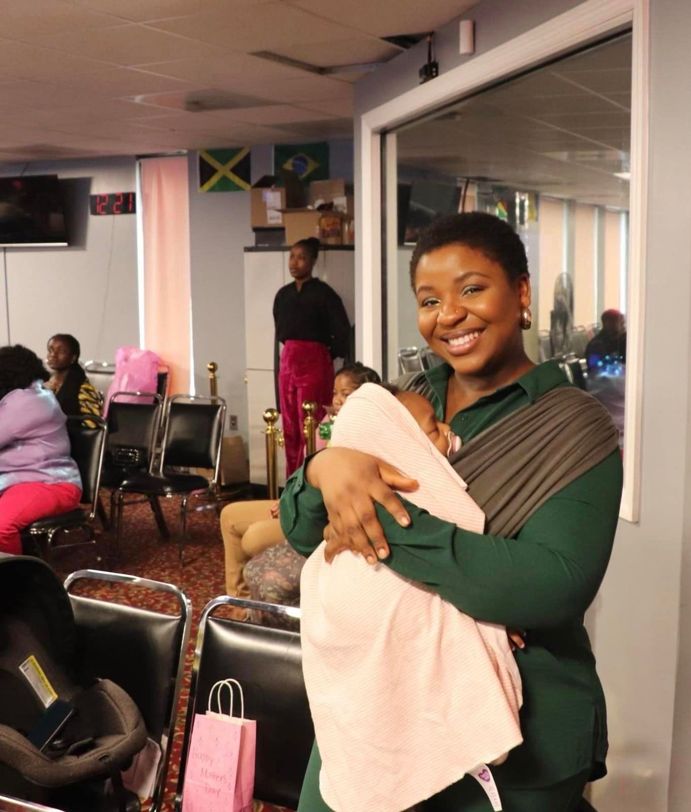 Woman in green shirt smiles while holding a baby in a carrier at an event.