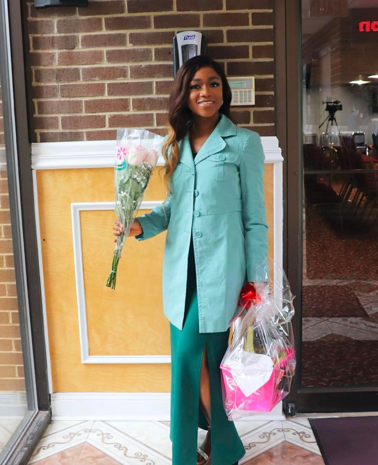 Woman in green suit holding flowers and gift, smiling outside a building.