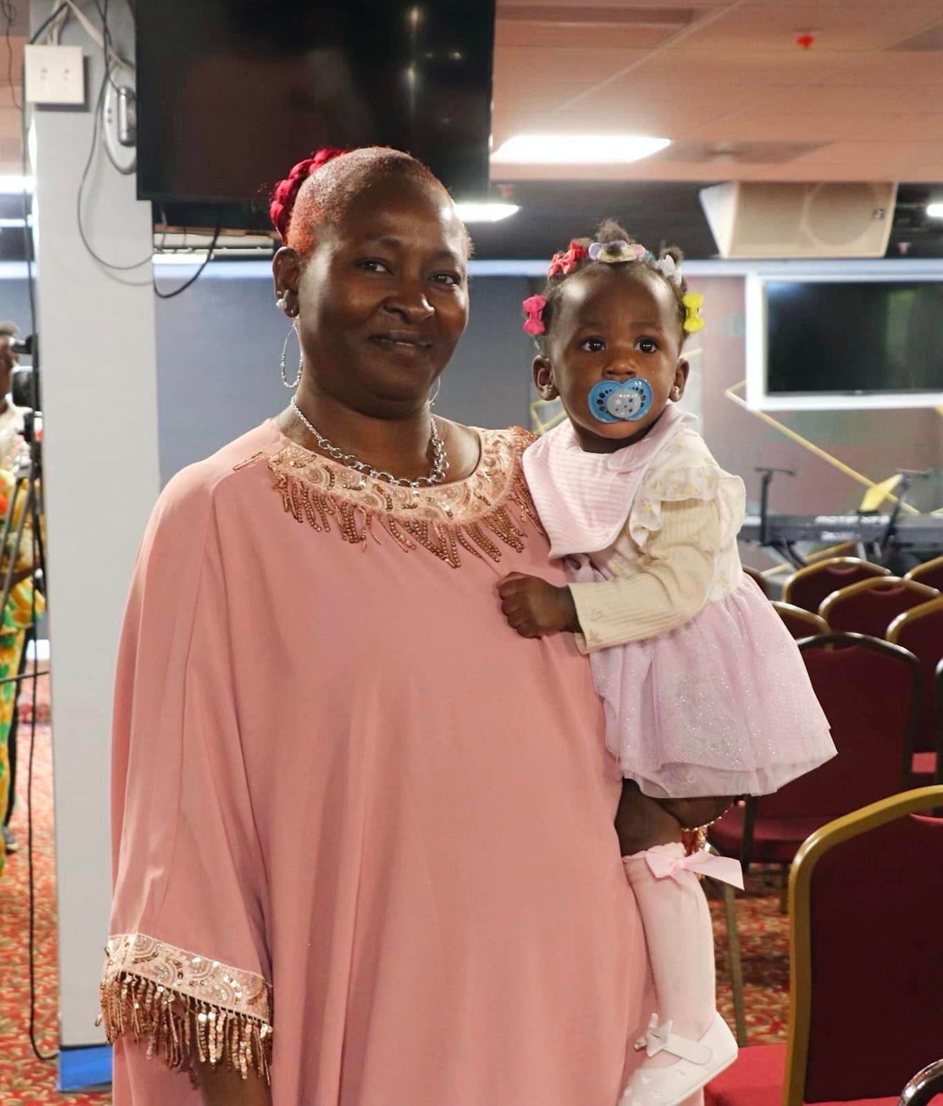Woman in pink dress holding a baby in a tutu, both smiling. Indoors with chairs.